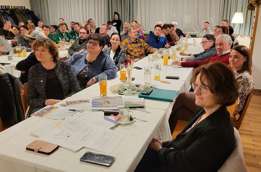 Eine Gruppe von Menschen sitzt an einem langen Tisch in einer Besprechung. Auf dem Tisch liegen Papiere, Stifte und Gläser mit Orangensaft.
