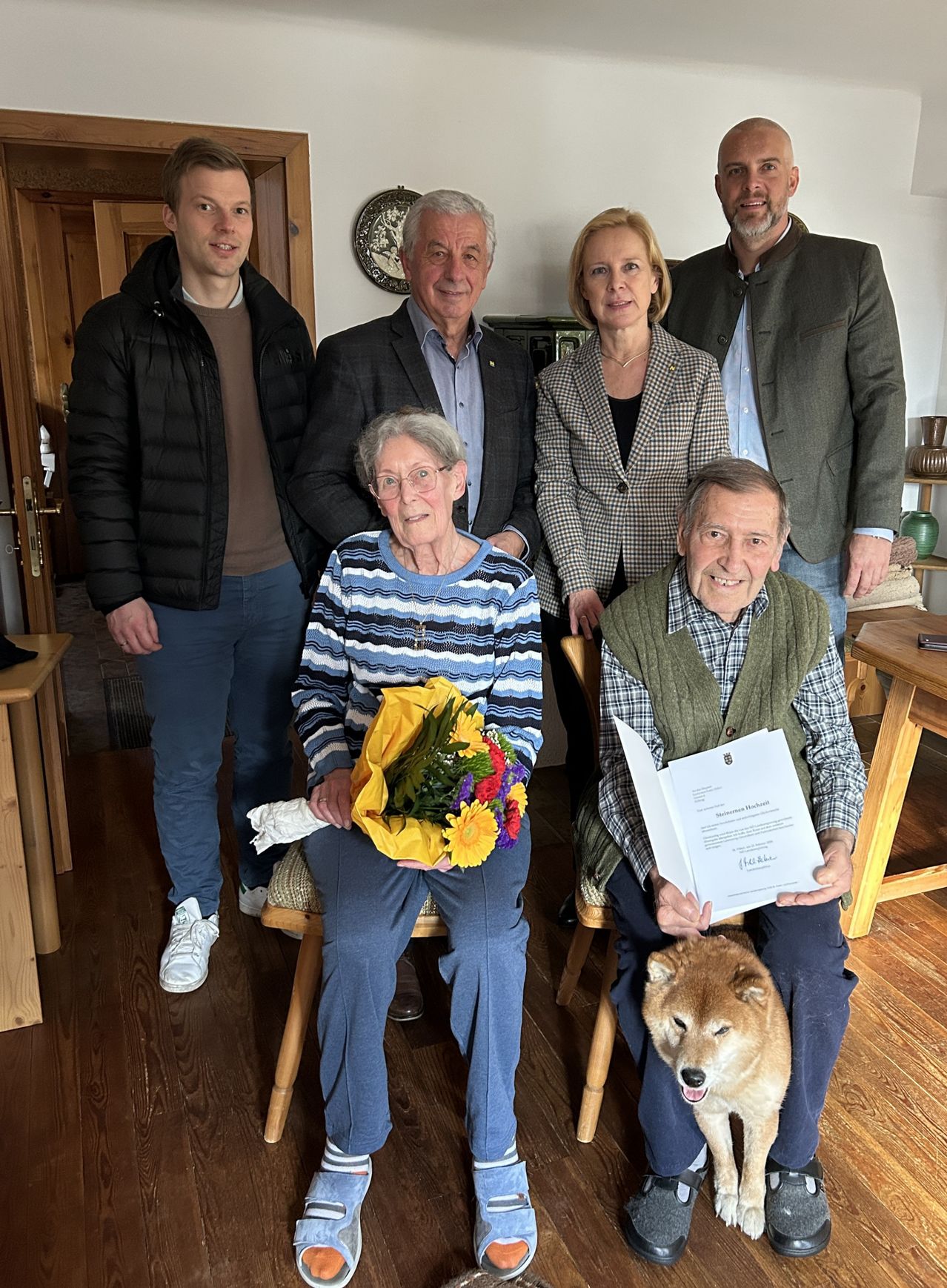 Eine Gruppe von Menschen in einem Raum mit Holzböden, die lächelnd für ein Foto posieren. Eine ältere Frau mit Brille hält einen Blumenstrauß. Der ältere Mann neben ihr hält ein Zertifikat. Die anderen stehen und sitzen. Dahinter befinden sich Regale mit verschiedenen Gegenständen.