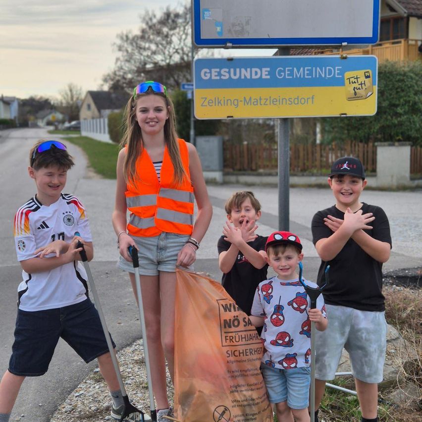 Eine Gruppe von Kindern steht auf einer Straße. Ein Mädchen in einer orangefarbenen Weste hält einen Müllsack, während andere Stöcke halten. Dahinter befindet sich ein Schild für Zelking-Matzleinsdorf.