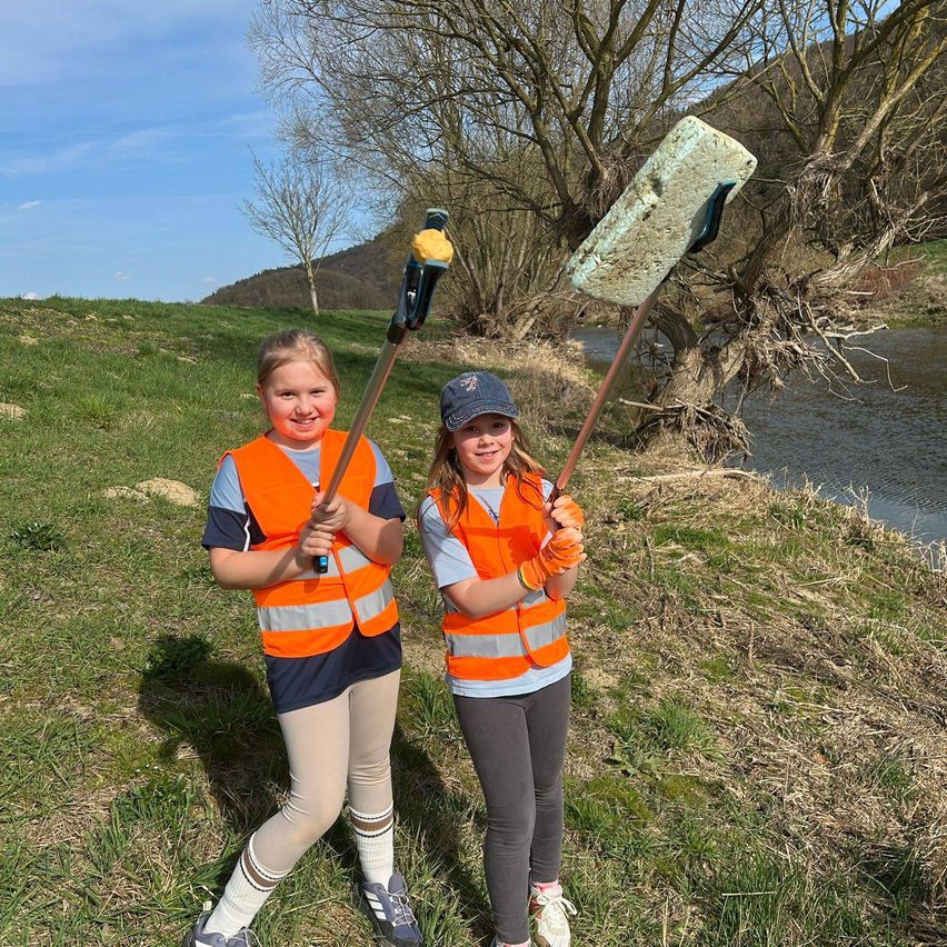 Zwei junge Mädchen in orangenen Westen halten lange Werkzeuge in der Hand und stehen auf Gras in der Nähe eines Flusses und lächeln. Bäume und Berge sind im Hintergrund.
