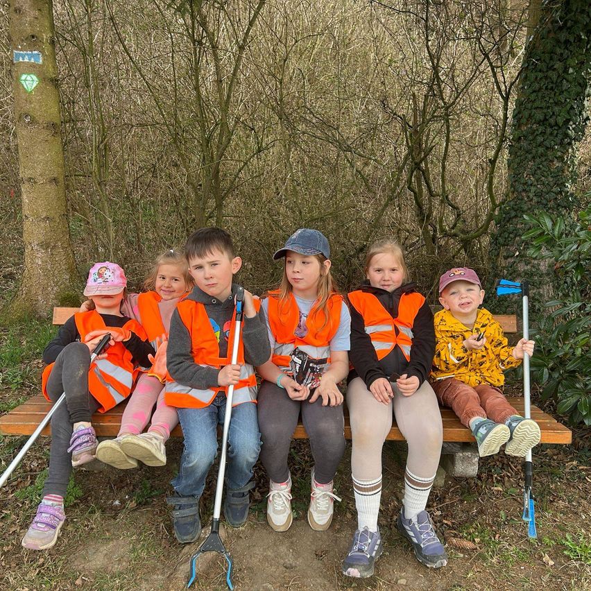 Eine Gruppe von Kindern, die orange Westen tragen, sitzt auf einer Bank im Wald und hält Stöcke. Dahinter sind Bäume zu sehen, von denen einer ein Diamantsymbol hat.
