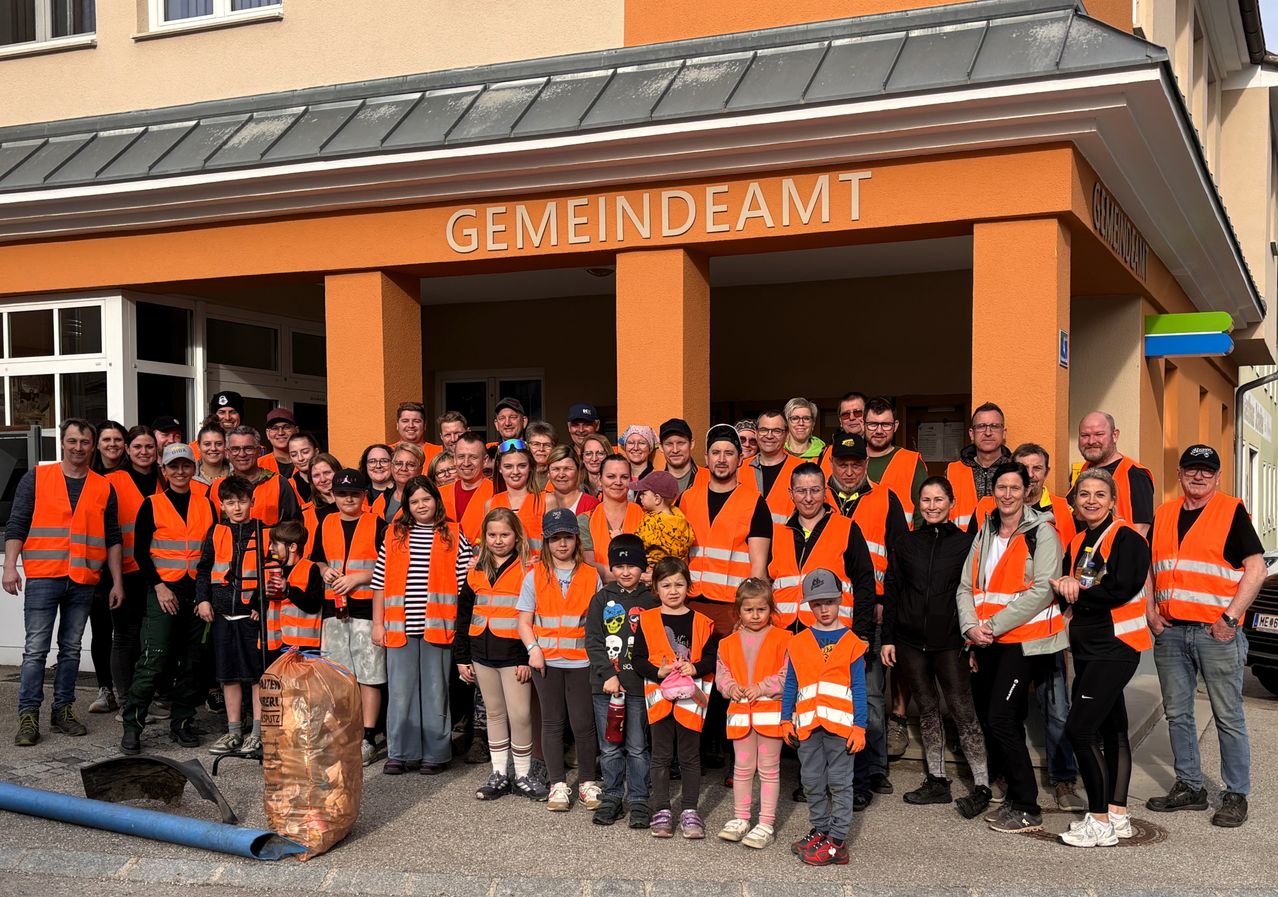 Eine Gruppe von Menschen in orangefarbenen Westen posiert für ein Foto vor einem Gebäude mit der Aufschrift Gemeindeamt. Einige halten Taschen und Flaschen, und ein Müllsack liegt auf dem Boden.