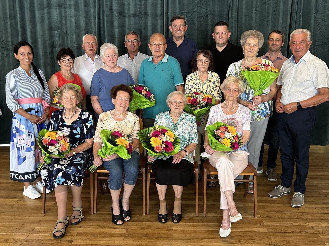 Eine Gruppe älterer Personen, sowohl Männer als auch Frauen, sind für ein Foto versammelt. Sie sitzen und stehen und halten Blumensträuße. Der Hintergrund scheint drinnen zu sein mit einem grünen Vorhang.