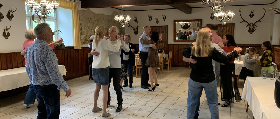 Eine Gruppe von Menschen tanzt in einem Raum mit Holzbalken, Kronleuchtern und einem Spiegel. Einige sind zu zweit, andere stehen herum. Tische und Stühle sind aufgestellt, und es gibt eine Wand mit einem Wandbild und einem Fenster.