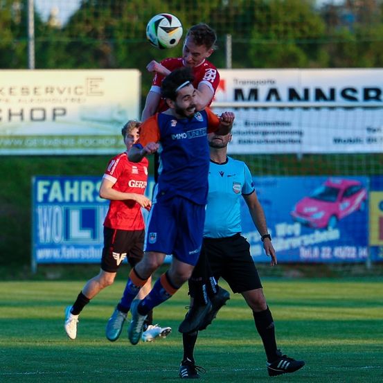 Ein Fußballspieler springt, um den Ball zu köpfen, während zwei Gegner versuchen, ihn zu blockieren, auf einem Feld mit Zaun und Bannern dahinter.