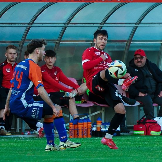 Ein Fußballspieler tritt den Ball, während ein anderer Spieler und Zuschauer zusehen. Der Spieler in Rot trägt rote Schuhe. Im Hintergrund befinden sich Flaschen und eine Tasche.
