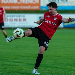Ein Fußballspieler in Rot tritt einen Ball auf einem grünen Feld. Ein anderer Spieler steht im Hintergrund in der Nähe eines Werbebanners.