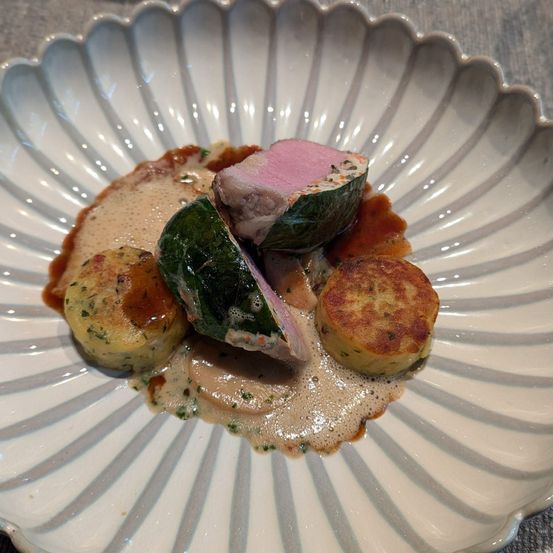Ein weißer Teller mit einem Gericht, das ein rosa Fleisch in grünen Blättern gewickelt, begleitet von kleinen, gebräunten Kartoffelpuffern und einer cremigen Sauce, zeigt.