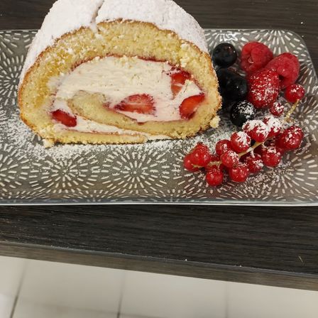 Ein gerollter Kuchen mit Erdbeer- und Sahnefüllung, bestreut mit Puderzucker, begleitet von Beeren auf einem dekorativen Teller.