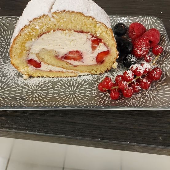 Ein gerollter Kuchen mit Erdbeer- und Sahnefüllung, bestreut mit Puderzucker, begleitet von Beeren auf einem dekorativen Teller.