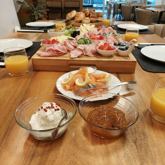 Ein Holztisch mit Tellern, Gläsern Orangensaft und einer Auswahl an Speisen wie Brot, Fleisch, Gemüse und Joghurt in Schalen.