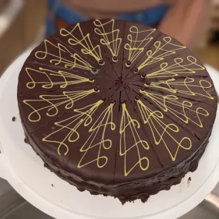 Ein Schokoladenkuchen mit gelben Wirbeln auf einem weißen Teller, die Hand von jemandem ist im Hintergrund sichtbar.