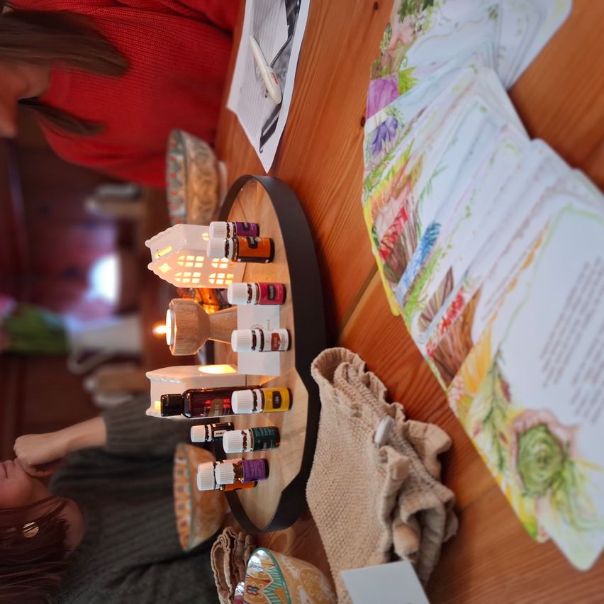 A woman in a red sweater sits at a wooden table with various essential oils and candles on a tray. Papers and a cloth are nearby.