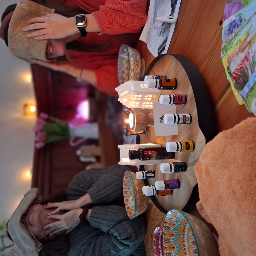 Two people sit around a table with several bottles of essential oils, a candle, and a small lantern. The woman on the left wears a hat and a watch, and the room is softly lit.
