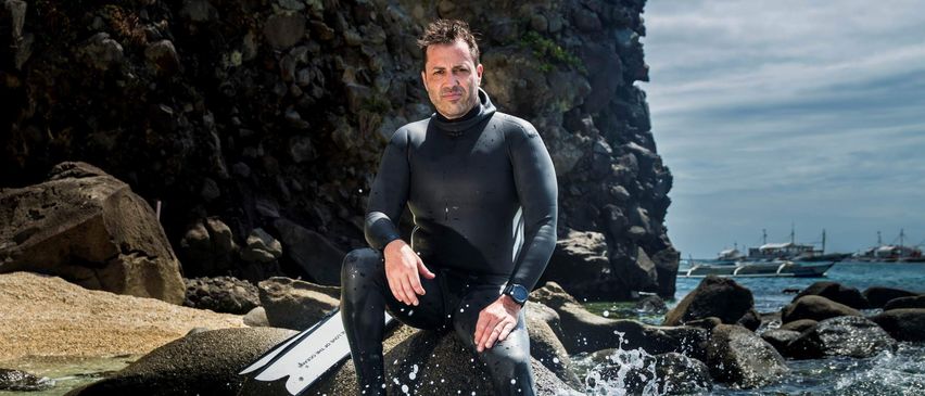 Ein Mann in einem schwarzen Neoprenanzug steht auf einem Felsen im Wasser, in der Nähe des Ufers.