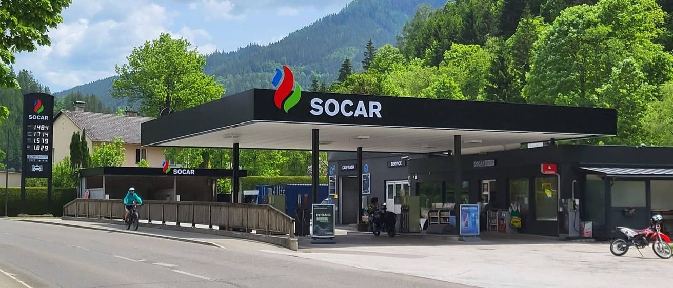 Eine SOCAR-Tankstelle mit schwarzem Vordach und einem Schild darüber. Eine Person fährt in der Nähe der Tankstelle mit dem Fahrrad.