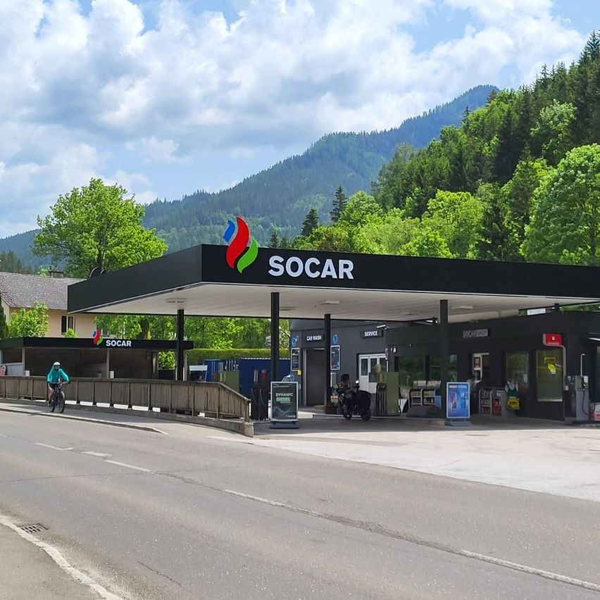Eine SOCAR-Tankstelle mit schwarzem Vordach und Berg im Hintergrund. Eine Person fährt ein Fahrrad in der Nähe des Eingangs.