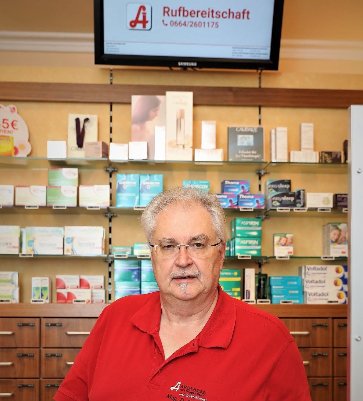 Ein älterer Mann mit Brille steht vor einer Apothekentheke, hinter der verschiedene Medikamente und Produkte ausgestellt sind.