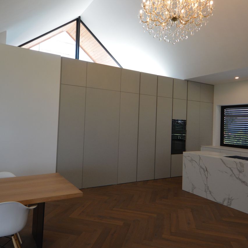 Innenansicht eines modernen Esszimmers mit Holzboden und weißen Wänden, ausgestattet mit einer großen Kristallkronleuchter, einem Kamin und einer Kücheninsel.