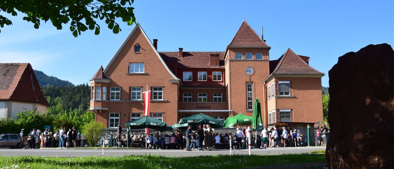 Ein großes braunes Gebäude mit einer Uhr und einer rot-weißen Fahne, umgeben von grünen Sonnenschirmen und Menschen, mit einem Grasbereich und Bäumen im Hintergrund.