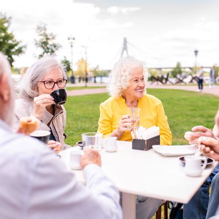 Mehrere ältere Menschen sitzen um einen Tisch in einem Park und genießen Getränke und Essen. Eine Frau lächelt, während sie aus einem Becher trinkt. Eine andere Frau hält ein Glas. Sie scheinen in ein Gespräch vertieft zu sein.