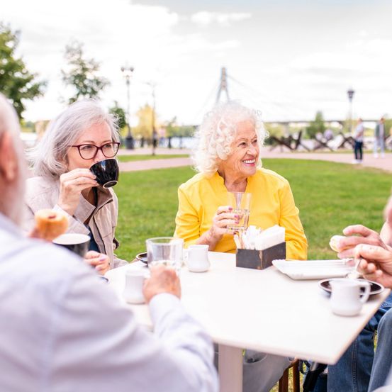 Mehrere ältere Menschen sitzen um einen Tisch in einem Park und genießen Getränke und Essen. Eine Frau lächelt, während sie aus einem Becher trinkt. Eine andere Frau hält ein Glas. Sie scheinen in ein Gespräch vertieft zu sein.