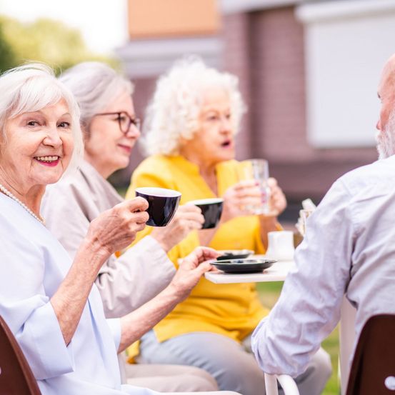 Eine Gruppe älterer Frauen und ein Mann sitzen um einen Tisch herum, halten Tassen und unterhalten sich. Die Frau vorne hält eine schwarze Tasse. Im Hintergrund ist eine verschwommene Ansicht eines Hauses zu sehen.