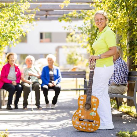 Eine Frau steht draußen mit einer Gitarre in der Hand und lächelt. Hinter ihr sitzen drei ältere Frauen auf einer Bank, eine mit einem Stock. Sie befinden sich in einem Park mit Bäumen und einem Gebäude im Hintergrund.
