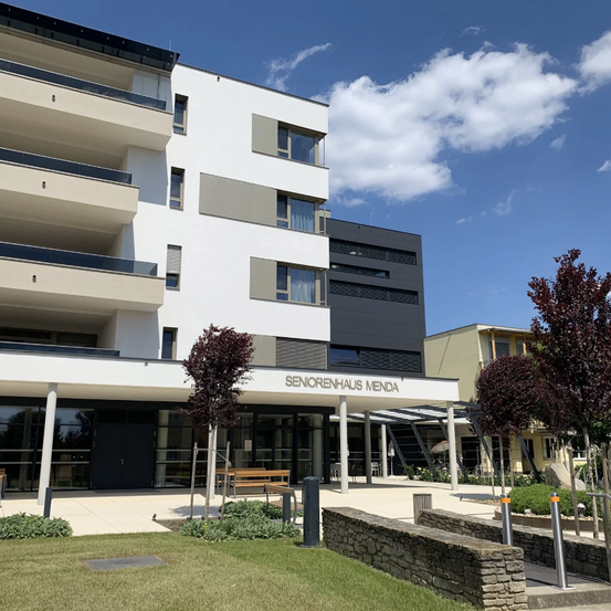 Das Seniorenheim Mend Gebäude ist ein modernes mehrstöckiges Bauwerk mit Glasfenstern, einem großen Eingang und einem kleinen Garten mit Bäumen und Bänken.