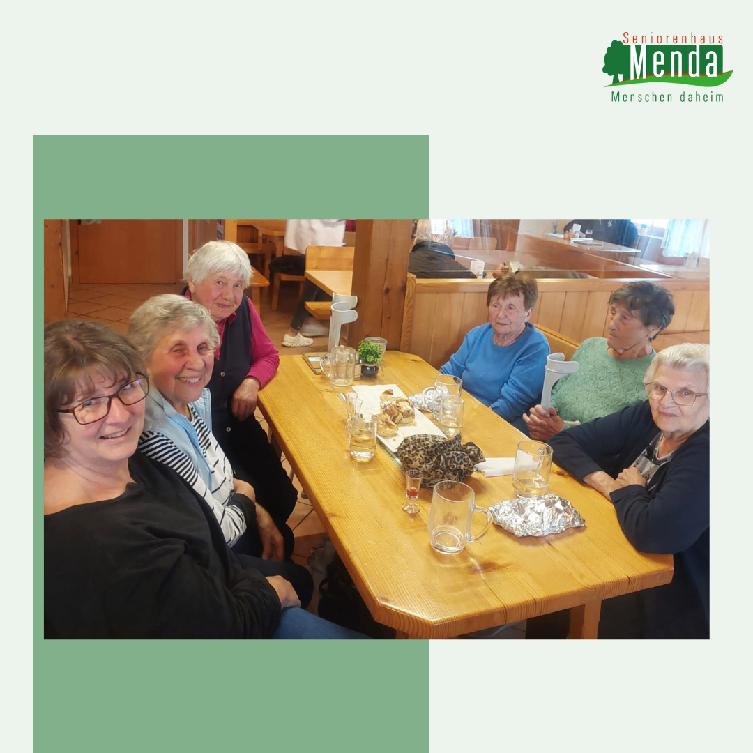 Eine Gruppe älterer Frauen sitzt lächelnd um einen Holztisch in einem Restaurant. Auf dem Tisch stehen Essen, Gläser und eine Karaffe. Hinter ihnen sitzt eine Person auf einem Stuhl. Ein Logo mit den Worten 'Seniorenhaus Menda' befindet sich in der oberen rechten Ecke.