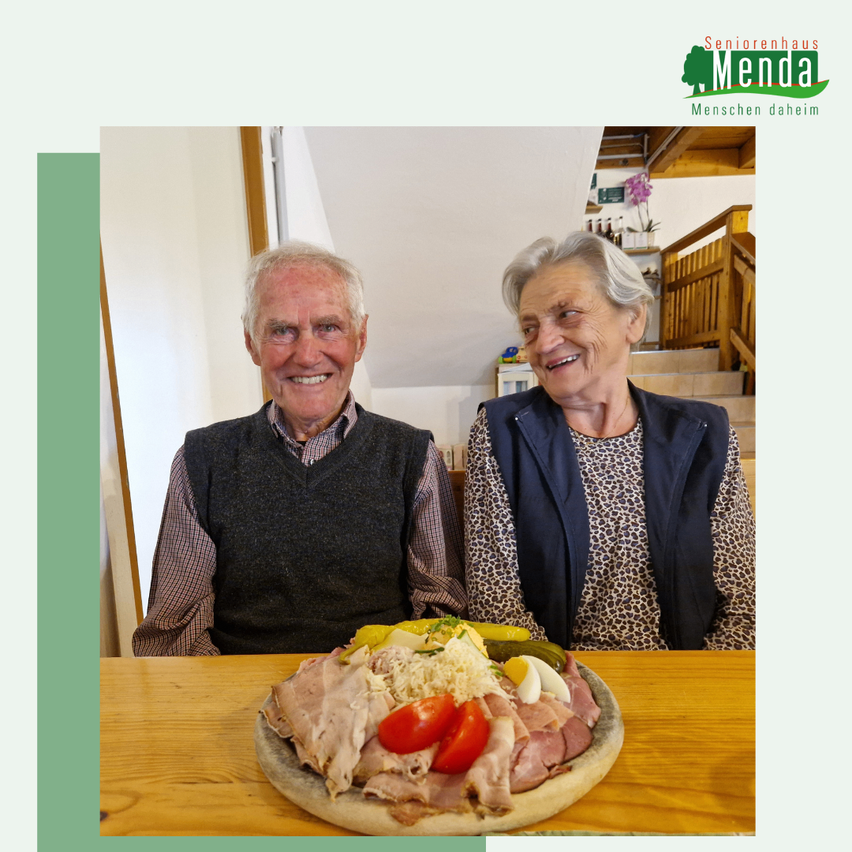 Zwei ältere Menschen sitzen neben einem Tisch mit einem Fleischplatten. Der Mann lächelt, während die Frau lächelt und in die Kamera schaut.