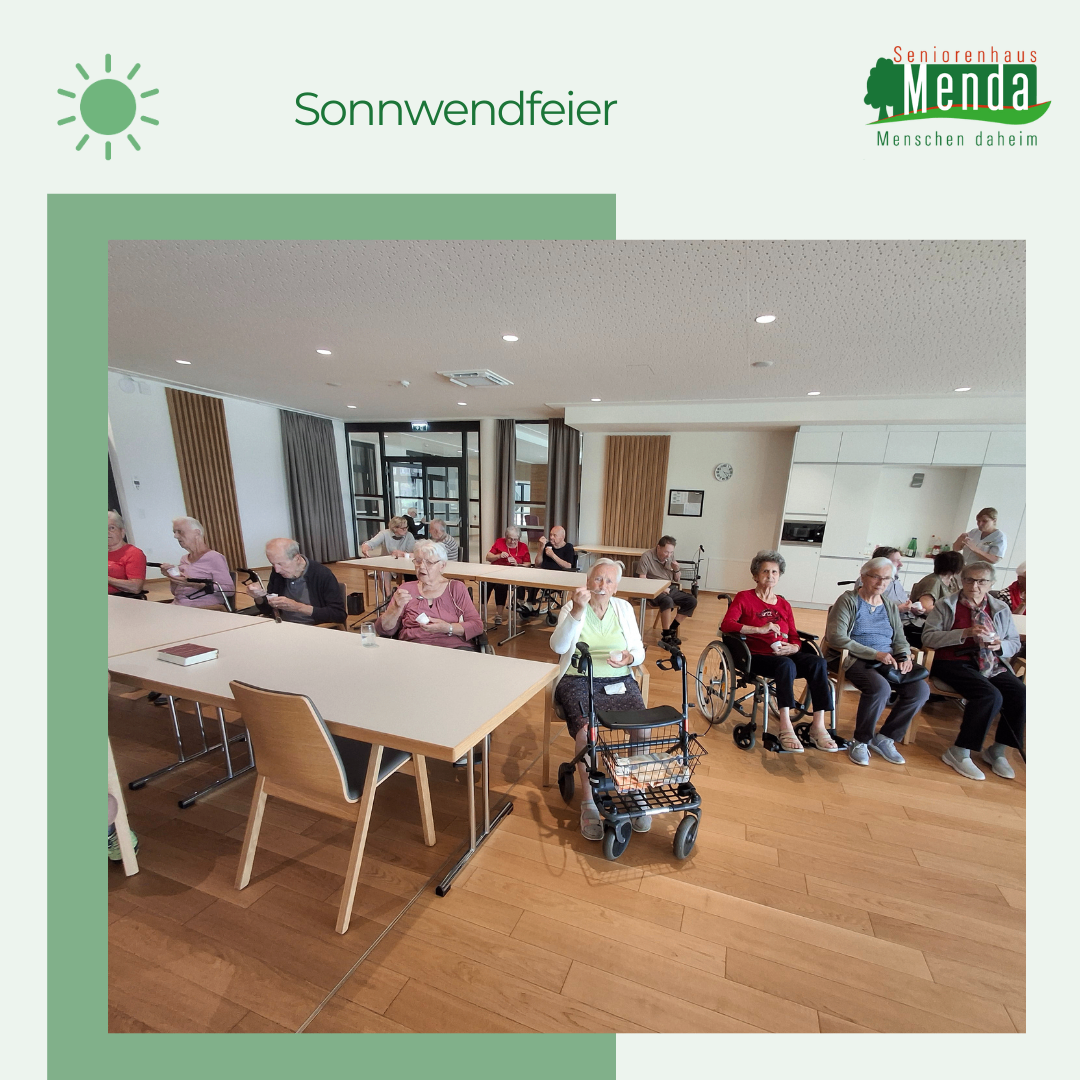 Eine Gruppe älterer Menschen in Rollstühlen und Stühlen sitzt in einem Raum mit Holzboden und weißen Wänden. Eine Frau in einem Rollstuhl mit Gehhilfe steht vorne. Es gibt Tische mit Büchern und ein Sonnensymbol in der oberen linken Ecke.