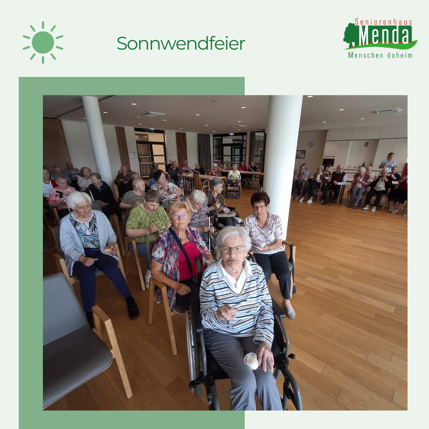 Eine Gruppe älterer Menschen sitzt in Stühlen, einige in Rollstühlen, in einem Raum. Eine Frau mit Brille und gestreifter Bluse hält einen Becher. Ein Logo lautet 'Seniorenhaus Menda'.