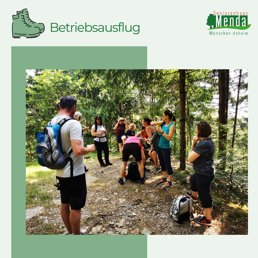 Eine Gruppe von Menschen wandert im Wald, einige mit Rucksäcken, andere ruhen sich aus, eine Frau vorne spricht.