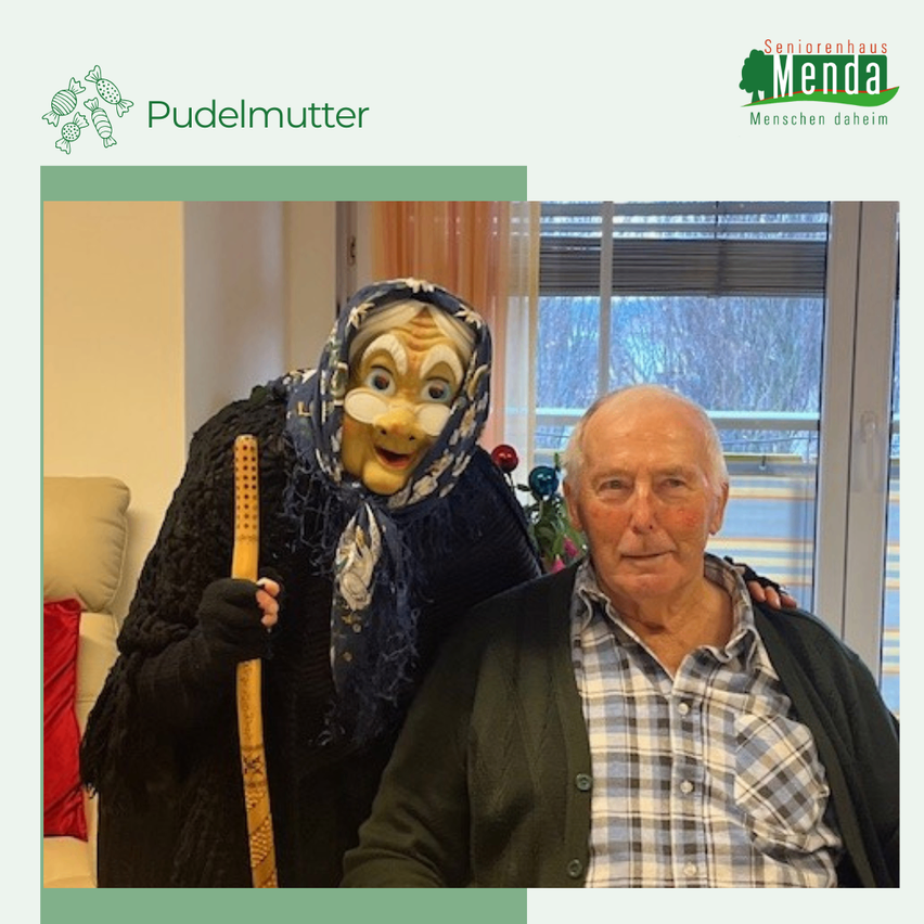 Ein älterer Mann posiert mit einer als Hexe verkleideten Person, die einen Stock hält. Hinter ihnen ist ein Fenster mit Jalousien.