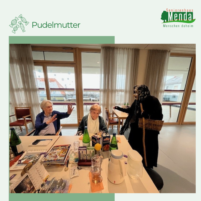 Eine Gruppe älterer Menschen sitzt um einen Tisch herum, einer trägt eine Maske. Der Tisch hat Flaschen, Bücher und andere Gegenstände. Sie befinden sich in einem Raum mit Glasfenstern.