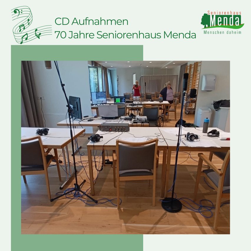 Ein Innenraum eines Aufnahmestudios mit mehreren Schreibtischen, Mikrofonen und Stühlen. Der Raum hat Holzböden und weiße Wände. Im Hintergrund sind Personen zu sehen.