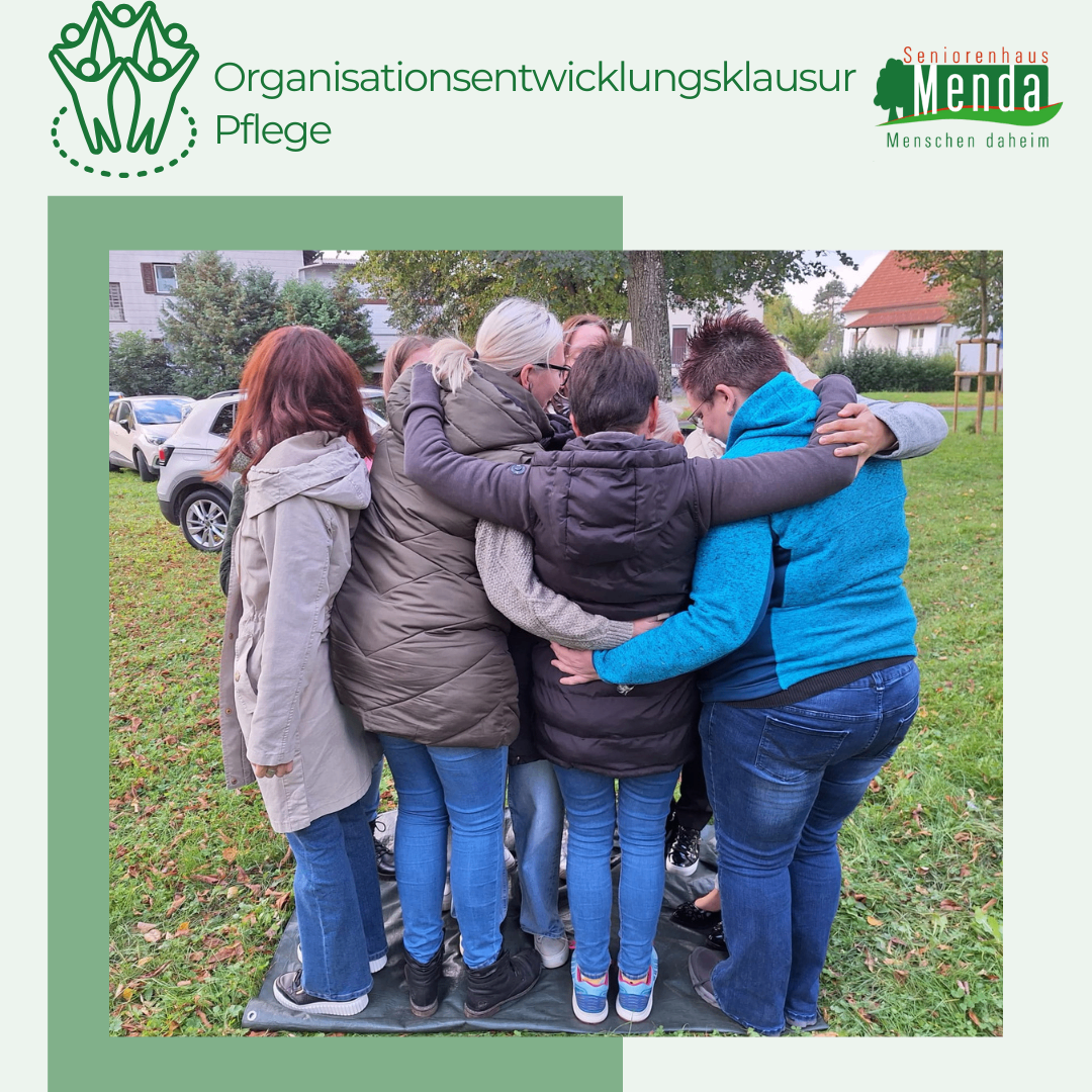Fünf Frauen stehen eng beieinander in einem Park und teilen einen warmen Moment. Sie sind von Gras und einigen Gebäuden umgeben.