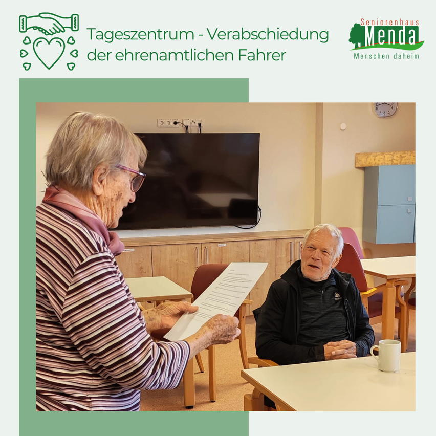 Eine ältere Frau mit einem Papier in der Hand steht neben einem älteren Mann, der an einem Tisch sitzt. Hinter ihnen ist ein Fernseher an der Wand montiert. Die Umgebung scheint ein Gemeindezentrum zu sein.