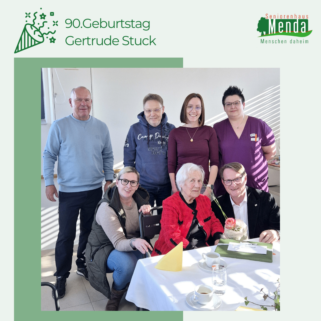 Ein Gruppenfoto von sechs Personen, die Gertrude Stucks 90. Geburtstag in einem Raum mit Tisch, Tassen und einer Blume feiern.