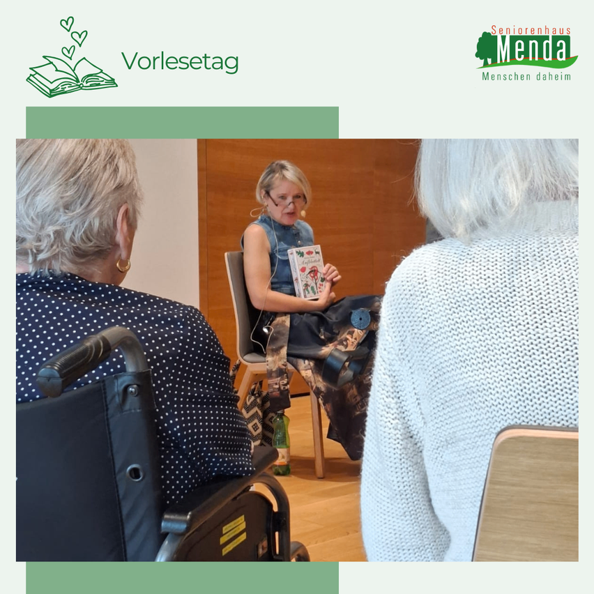 Eine Veranstaltung, bei der eine Frau ein Buch für eine Gruppe älterer Menschen in einem Raum vorliest. Eine Frau im Rollstuhl und eine andere in einem Stuhl lauschen aufmerksam zu.