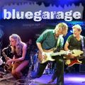 Eine Band tritt auf der Bühne auf, mit einer Frau, die Saxophon spielt, und drei Männern, die Gitarre spielen. Der Hintergrund ist lila und das Wort 'bluegarage' ist prominent angezeigt.