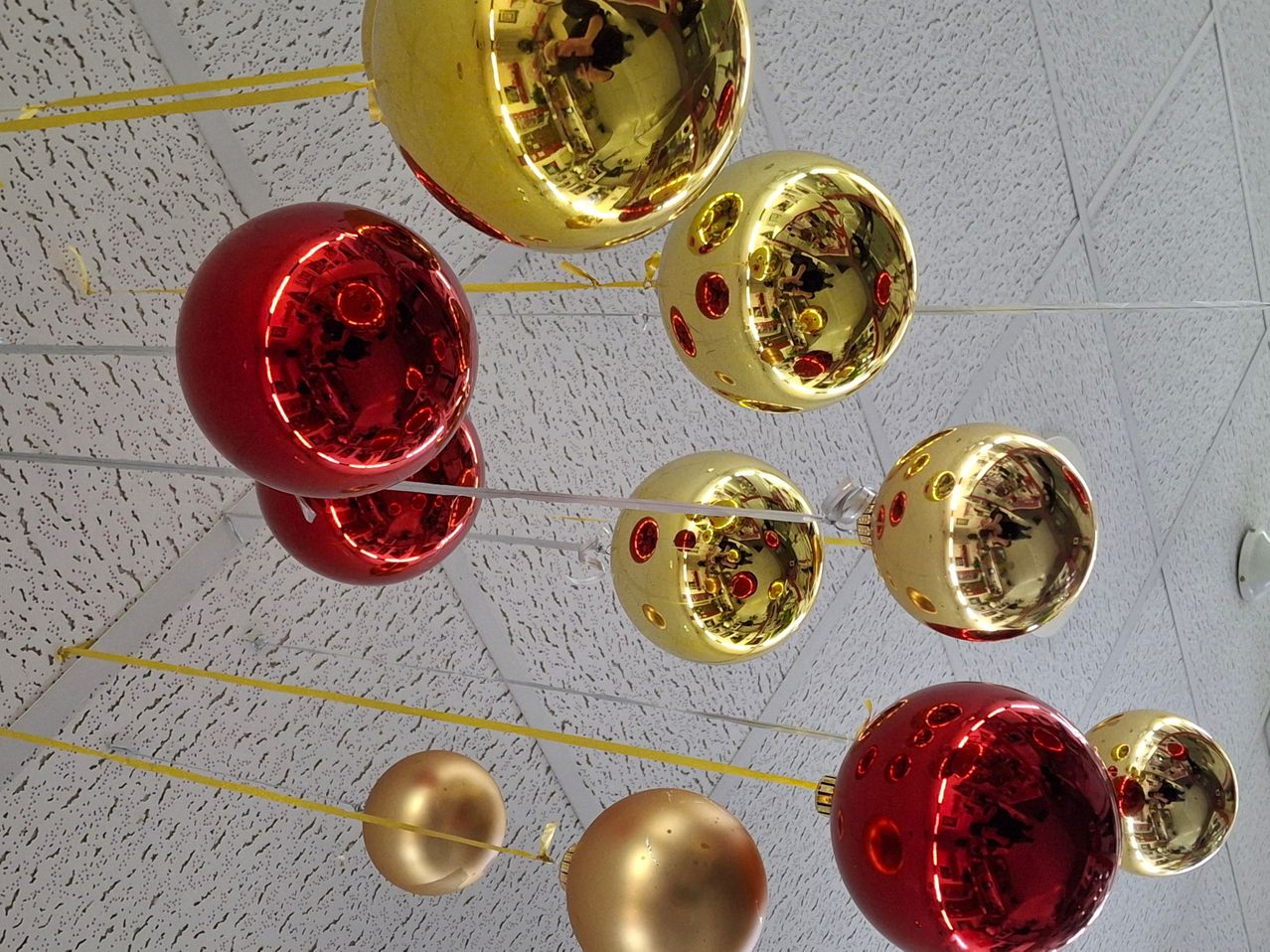 Eine Sammlung kugelförmiger Ornamente, abwechselnd rot und gold, hängt von einer strukturierten Decke und reflektiert die Umgebung.