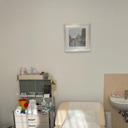 Ein Untersuchungsraum mit einem weißen Bett, einem Waschbecken und einem Schrank mit medizinischen Utensilien auf einem gefliesten Boden.