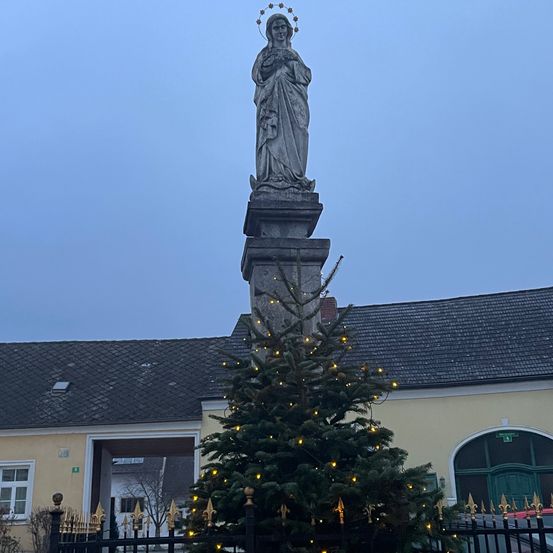 Eine Statue einer Frau mit Krone steht auf einem Steinpfeiler mit einem Weihnachtsbaum vor einem Gebäude.