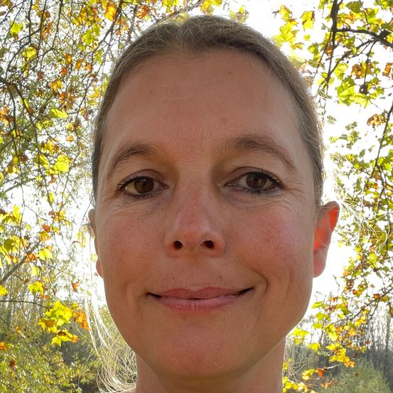 Eine Frau in einem grünen Kittel lächelt für die Kamera, mit Bäumen und Laub im Hintergrund.