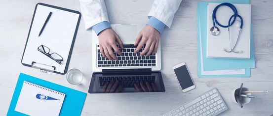 Eine Person in einem weißen Laborkittel tippt auf einem Laptop mit einem Mobiltelefon, Notizbuch und Brille auf einem Schreibtisch.