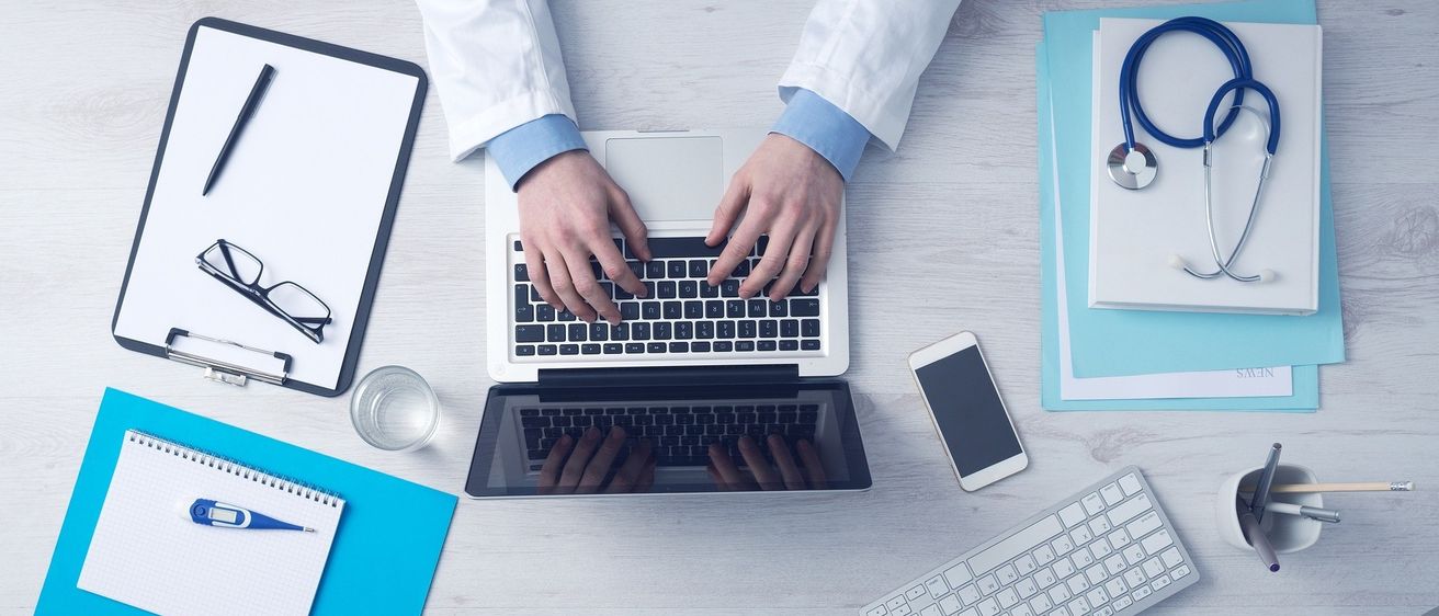 Eine Person in einem weißen Laborkittel tippt auf einem Laptop mit einem Mobiltelefon, Notizbuch und Brille auf einem Schreibtisch.
