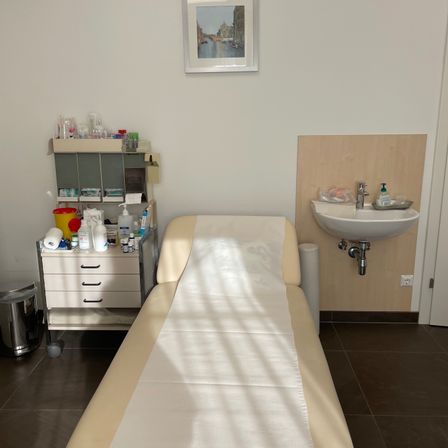 Ein Untersuchungsraum mit einem Bett, einem Waschbecken und einem Schrank mit verschiedenen medizinischen Utensilien. Ein gerahmtes Bild schmückt die Wand.