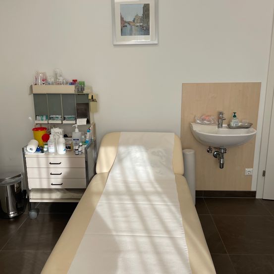 Ein Untersuchungsraum mit einem Bett, einem Waschbecken und einem Schrank mit verschiedenen medizinischen Utensilien. Ein gerahmtes Bild schmückt die Wand.